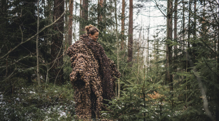 Nainen seisoo metsässä yllään kävyistä tehty viitta.