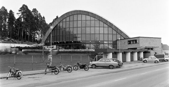 Mustavalkoisessa kuvassa näkymä Palomäenkadulta kohti uimahallin kaarirakennusta. Etualalla uimahallin edustalle pysäköitynä neljä moottoripyörää ja kaksi henkilöautoa. Autojen takaa näkyy hieman yhtä moottoripyörää. Uimahallin sisäänkäynnin edessä pysäköitynä kaksi henkilöautoa, pakettiauto sekä muutama polkupyörä. Vasemmalla näkyy hieman uimahallin ulkoallasta.