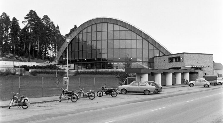 Mustavalkoisessa kuvassa näkymä Palomäenkadulta kohti uimahallin kaarirakennusta. Etualalla uimahallin edustalle pysäköitynä neljä moottoripyörää ja kaksi henkilöautoa. Autojen takaa näkyy hieman yhtä moottoripyörää. Uimahallin sisäänkäynnin edessä pysäköitynä kaksi henkilöautoa, pakettiauto sekä muutama polkupyörä. Vasemmalla näkyy hieman uimahallin ulkoallasta.