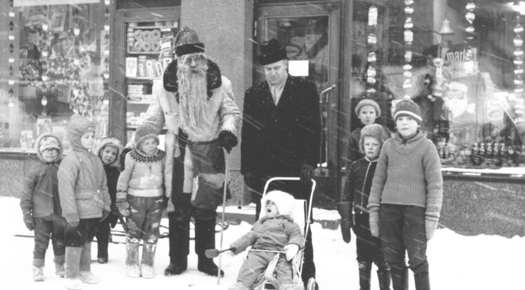 Joulupukki ja pankinjohtaja Paavo Koponen keskellä. Koponen pitää kiinni lastenrattaissa, joissa istuu hänen poikansa leikkilapio kädessään. Koposen oikealla puolella kolme poikaa talvivaatteissa. Joulupukin vieressä vasemmalla neljä lasta talvivaatteissa. Takana Voikan Herkun jouluiset näyteikkunat sekä sisäänkäynti liikkeeseen. Kuvassa sataa lunta ja maassa on lunta.