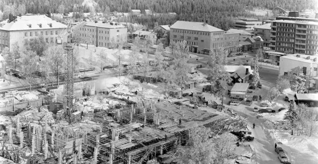 Etualalla näkyy uuden kaupungintalon rakennuksen perustuksia. Torninosturi rakennustyömaan keskellä sekä työmiehiä eri puolilla rakennustyömaata. Oikealla Torikadun varrella autoja pysäköitynä tien reunaan. Torikadun ja Kauppalankadun kulmauksessa oikealla Hotelli Kouvolan funkkisrakennuksen päätyä. Salaman talon takana näkyy Kouvolan Urheilupuiston uimahalli sekä Pikku-Palomäen kerrostaloja. Maassa on paljon lunta.