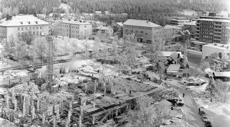 Etualalla näkyy uuden kaupungintalon rakennuksen perustuksia. Torninosturi rakennustyömaan keskellä sekä työmiehiä eri puolilla rakennustyömaata. Oikealla Torikadun varrella autoja pysäköitynä tien reunaan. Torikadun ja Kauppalankadun kulmauksessa oikealla Hotelli Kouvolan funkkisrakennuksen päätyä. Salaman talon takana näkyy Kouvolan Urheilupuiston uimahalli sekä Pikku-Palomäen kerrostaloja. Maassa on paljon lunta.