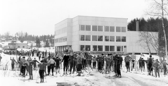 Etualalla paljon koululaisia hiihtovarusteissa kasvot kuvaan päin. Taustalla valkoinen, kuutiomainen Hirvelän koulun rakennus. Takana vasemmalla koulun lähellä sijainneita puisia asuinrakennuksia.