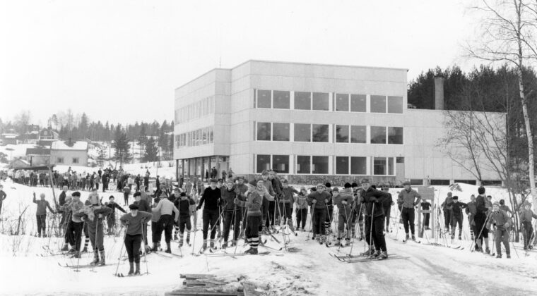 Etualalla paljon koululaisia hiihtovarusteissa kasvot kuvaan päin. Taustalla valkoinen, kuutiomainen Hirvelän koulun rakennus. Takana vasemmalla koulun lähellä sijainneita puisia asuinrakennuksia.